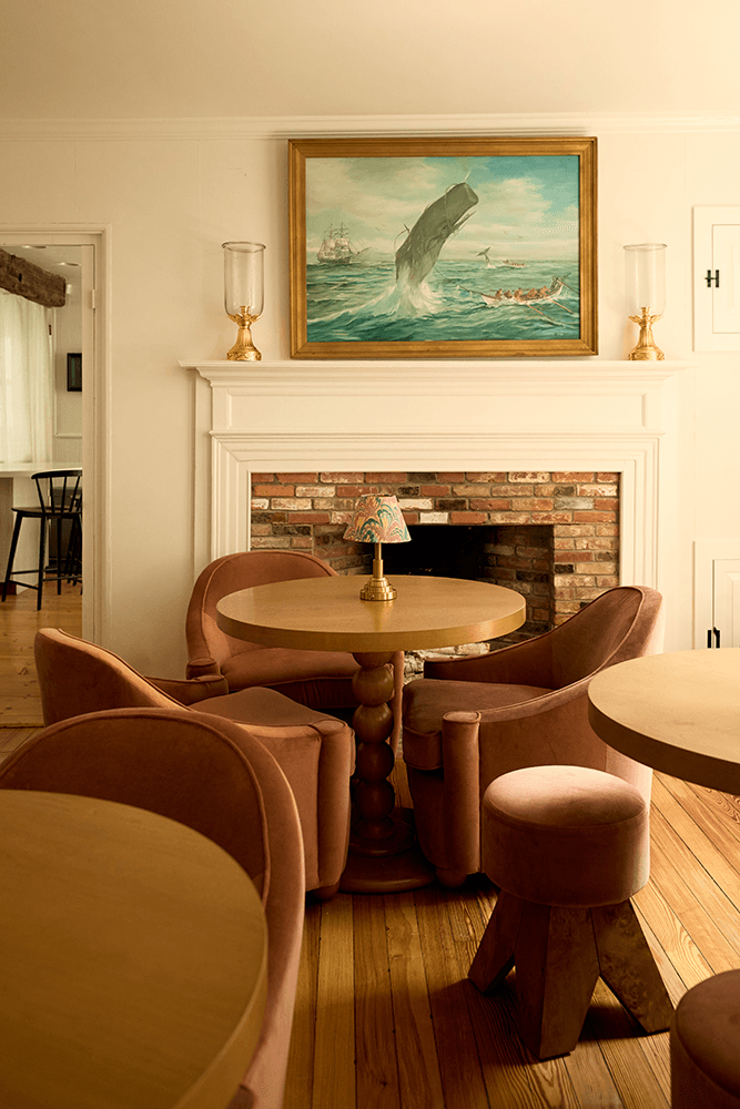 A cozy interior featuring pink furniture, a wooden table, and a whale painting above a brick fireplace.