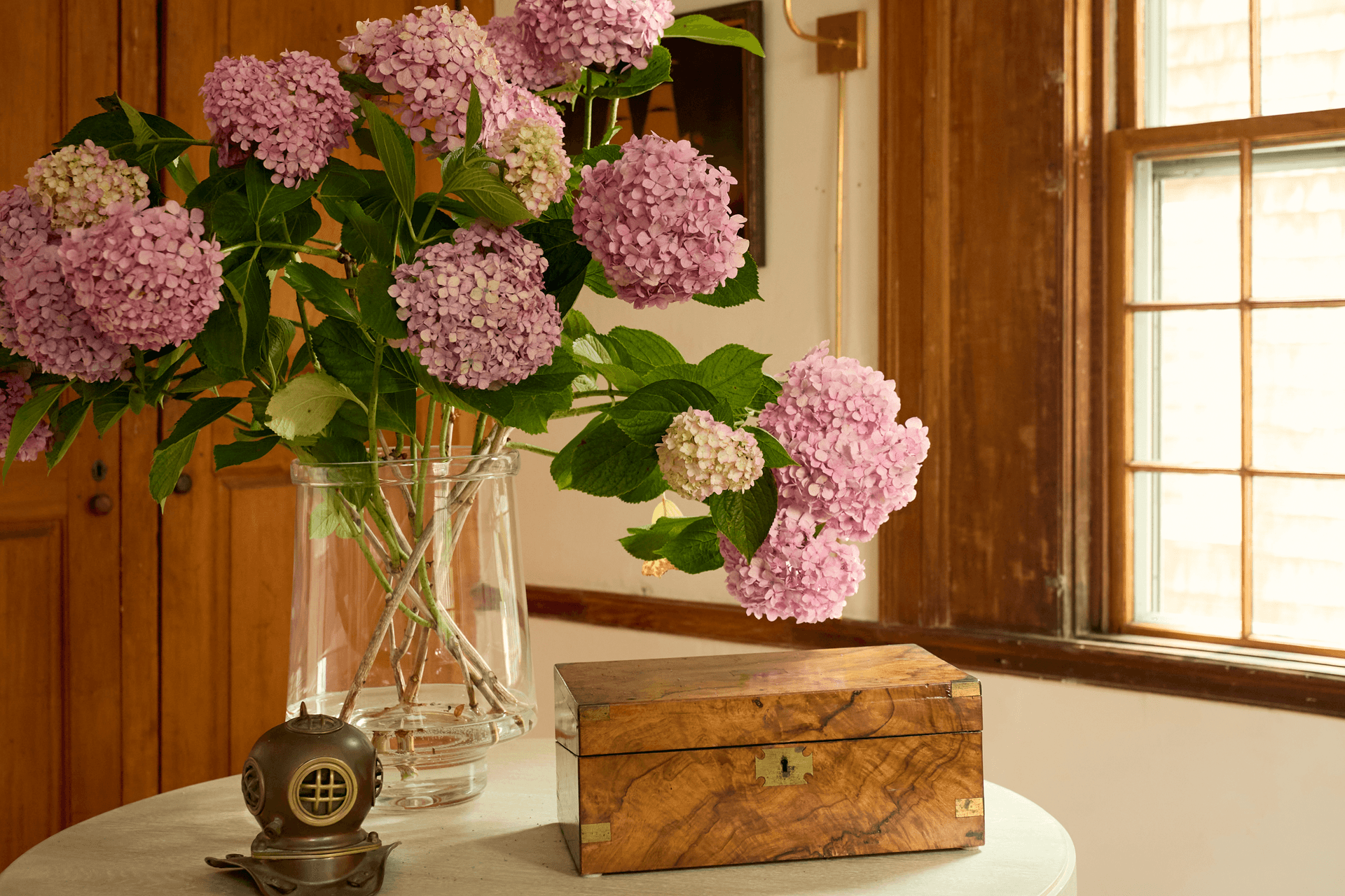 Purple hydrangeas in a glass vase, next to a wood box, on a round table in an entryway, in front of a brightly-lit window.
