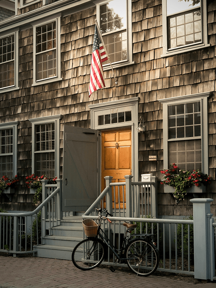 A rustic wooden house features a wooden door, a U.S. flag, and a bicycle parked on the porch.