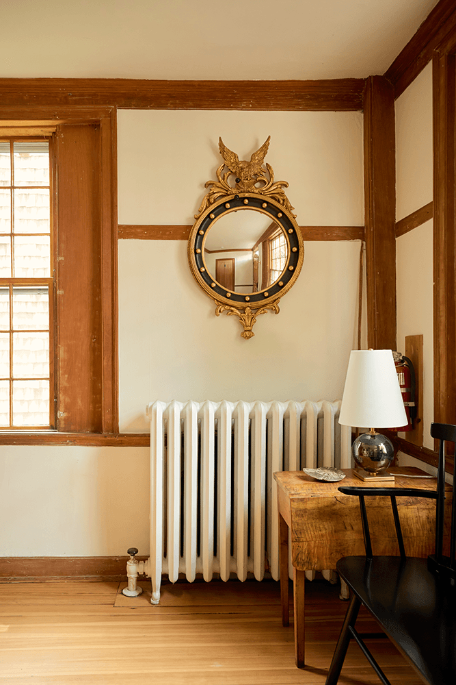 A vintage gold-framed mirror hangs above a wooden table and black chair, next to a white radiator in a room with wooden accents.