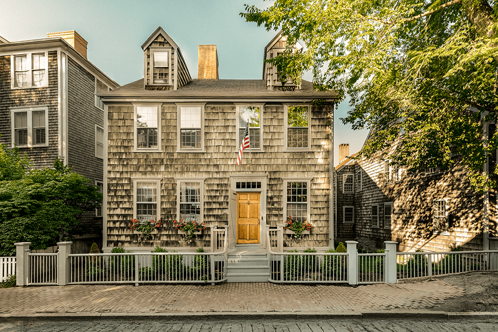A two-story wooden house with shingle siding, a central front door, an American flag, flower boxes, and a white picket fence, surrounded by trees.