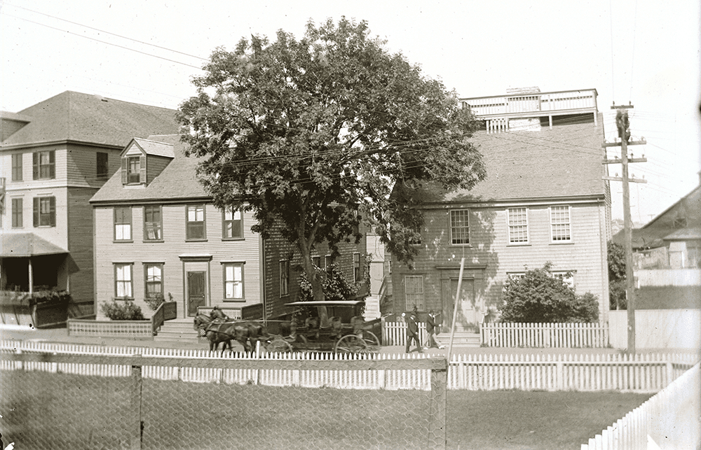 Black-and-white photo of a horse-drawn carriage on a street in front of wooden houses with a large tree and white picket fence; a person stands nearby.